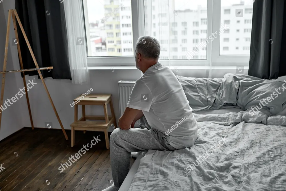 A man sitting on the edge of an unmade bed by the window